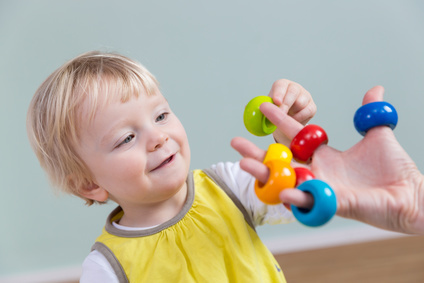 Kleines Kind lächelt und greift nach bunten Greifringen, während eine pädagogische Fachkraft die Ringe auf ihren Fingern hält; freundliche Betreuungssituation und spielerisches Üben feinmotorischer Fähigkeiten.