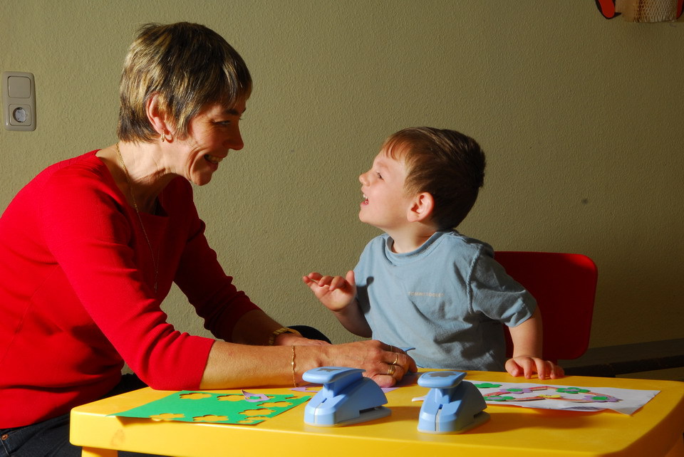 Eine lächelnde Pädagogin im roten Pullover und ein kleiner Junge im blauen T-Shirt blicken sich am Basteltisch glücklich an. Sie nutzen Motivstanzer für grünes Papier.