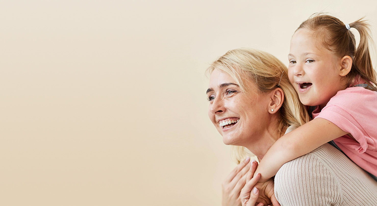 Eine lächelnde Mutter und ihre Tochter mit Down-Syndrom, die sie huckepack trägt. Beide schauen fröhlich zur Seite vor einem hellen Hintergrund.