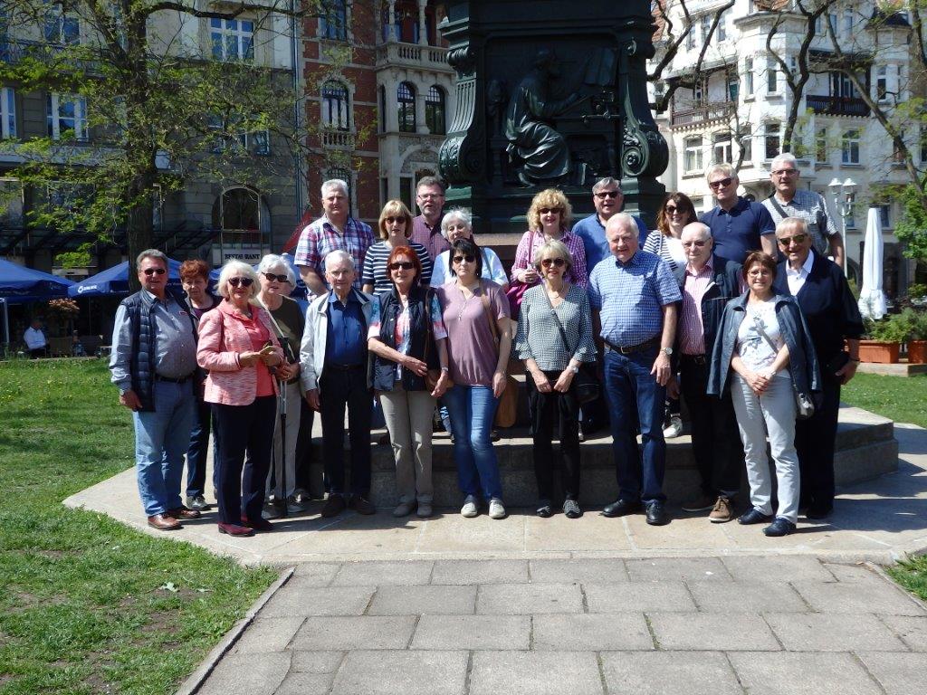Gruppenaufnahme von etwa 20 Seniorinnen und Senioren, die lächelnd auf einer Erhebung vor einem großen, dunklen Denkmal stehen. Im Hintergrund sind historische Gebäude und Sonnenschirme zu sehen.