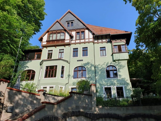 Repräsentative Außenaufnahme der Villa Karoline in Eisenach. Die Villa zeigt eine Mischung aus hellgrünem Putz und dekorativem dunklem Fachwerk im Giebelbereich. Mehrere Balkone und eine erhöhte Terrasse sind zu sehen, umrahmt von dichtem Grün. Eine Treppe und eine Stützmauer sind im Vordergrund.