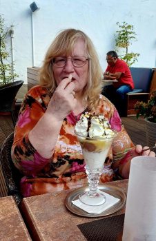 Eine Frau mit Brille isst in einer Betreuungssituation im Café ein großes Eisbecher-Dessert und hält einen Löffel, im Hintergrund sitzt eine weitere Person.