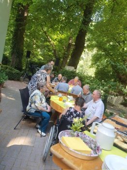 Mehrere Menschen mit Behinderung und Betreuungspersonen sitzen an einem langen Gartentisch unter Bäumen; eine pädagogische Fachkraft serviert Getränke, freundliche, entspannte Betreuungssituation bei einem gemeinsamen Essen.
