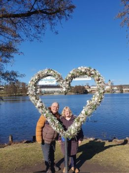 Zwei lächelnde Menschen, darunter eine pädagogische Fachkraft und eine Person mit Behinderung, stehen an einem See hinter einem großen, mit Blumen geschmückten Herzrahmen; im Hintergrund Stadtgebäude und blauer Himmel.