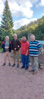 Vier Männer, darunter zwei ältere Herren und zwei Erwachsene mit sichtbarer Behinderung, stehen armseits nebeneinander lächelnd vor einem Wald- und Modellgebäudepanorama; die Stimmung wirkt freundlich und gemeinschaftlich.