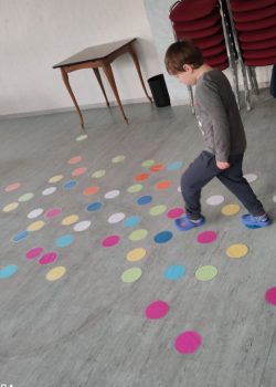 Kleiner Junge in Alltagskleidung läuft in einer Betreuungs- oder Spielsituation über bunte, auf dem Boden aufgelegte Papierkreise in einem Gruppenraum; stapelbare Stühle und ein Tisch im Hintergrund.