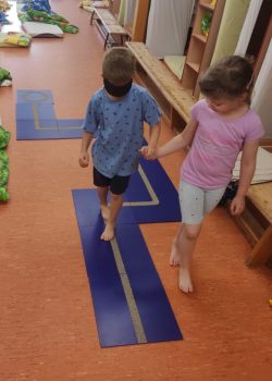 Zwei Kinder barfuß in einer Betreuungssituation: ein Kind mit Augenbinde tastet sich an einer blauen Bodenbahn entlang, gehalten an der Hand von einem weiteren Kind.