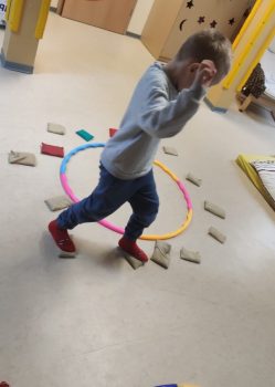 Kindesperson mit roten Hausschuhen balanciert in einer Betreuungs- bzw. Spielsituation über kleine Sandsäckchen um zwei bunte Reifen und wirkt konzentriert.