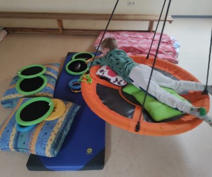 Ein Kind liegt bäuchlings auf einer runden Schaukel und greift nach Motorikringen auf Kissen; pädagogische Betreuungssituation in einem Innenraum mit Spiel- und Fördermaterial.