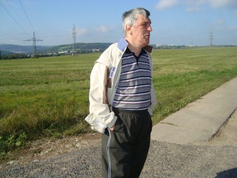 Mann mittleren Alters, vermutlich Mensch mit Behinderung, steht mit Händen in den Taschen am Wegesrand und blickt in ein weites Feld; ländliche Betreuungssituation bei Tageslicht.