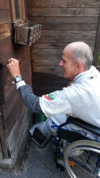 Mann im Rollstuhl nimmt mit Schraubenzieher an einem Holzanbau Reparaturarbeiten vor; freundliche, konzentrierte Betreuungssituation im Freien.