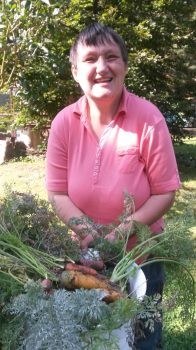 Frau in pinkem Polohemd hält lächelnd einen Eimer mit frisch geernteten, erdigen Möhren im Garten; Betreuungssituation im Freien, freundliche Stimmung.