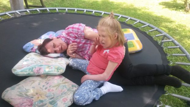 Pädagogische Fachkraft liegt auf einem Trampolin mit Kopfkissen und hält eine sitzende junge Person mit Behinderung aneinandergelehnt, beide im Freien und in einer ruhigen Betreuungssituation.