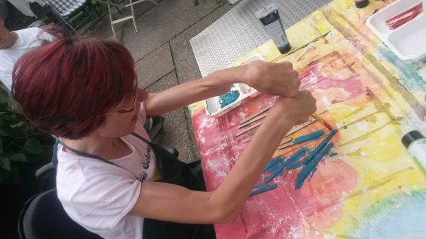 Frau mit rotem Haar und Brille sitzt an einem bemalten Tisch und bemalt mit Pinsel mehrere Holzstäbchen blau; Szene wirkt wie eine betreute künstlerische Beschäftigung.