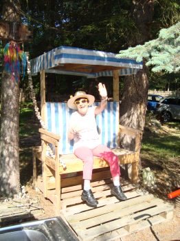 Frau mit Sonnenhut und gelber Sonnenbrille sitzt lächelnd auf einer Holzbank mit blau-weißer Markise und winkt in einer sonnigen Betreuungssituation im Freien.