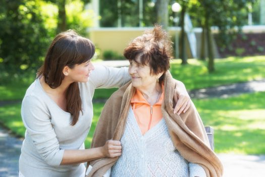 Junge pädagogische Fachkraft legt fürsorglich einen Mantel über eine ältere Frau mit Behinderung und beide lächeln in einer entspannten Betreuungssituation im Park.
