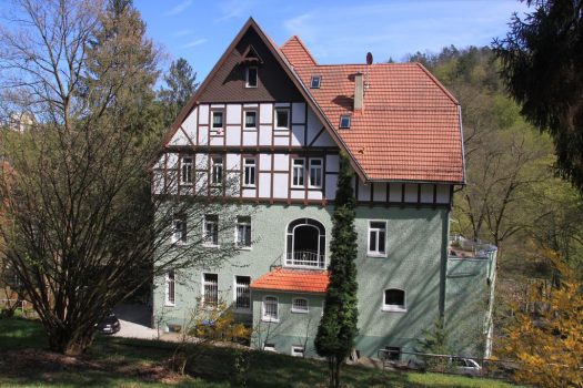 Dreigeschossiges Fachwerkhaus mit grün verputztem Erdgeschoss und rotem Satteldach in parkähnlicher Umgebung; Bäume und Sträucher umgeben das Gebäude, das nach außen als Einrichtung oder Wohnhaus wirkt.